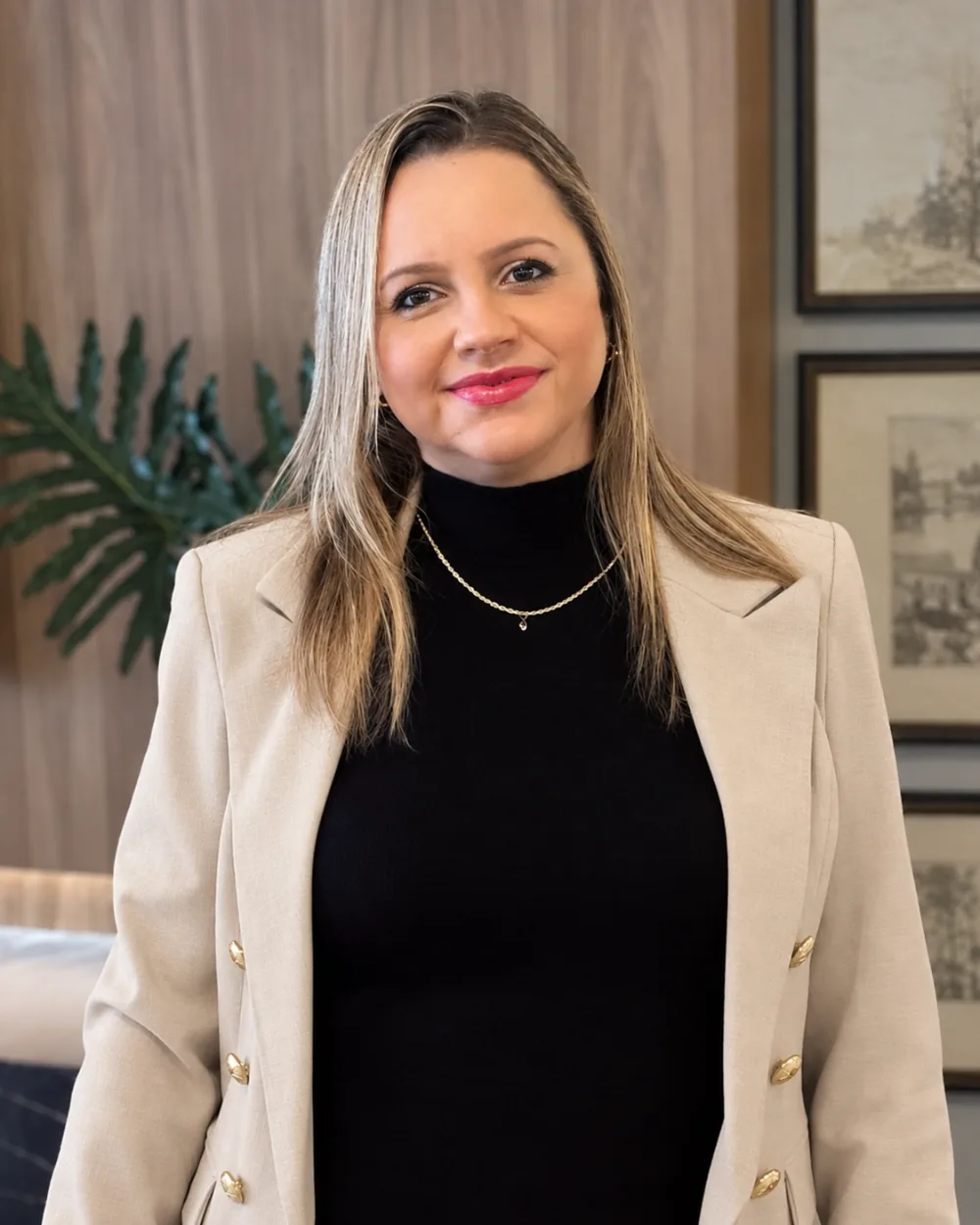 Daniele Brandão de blazer bege e blusa preta, sorrindo, diante de uma parede de madeira com quadros